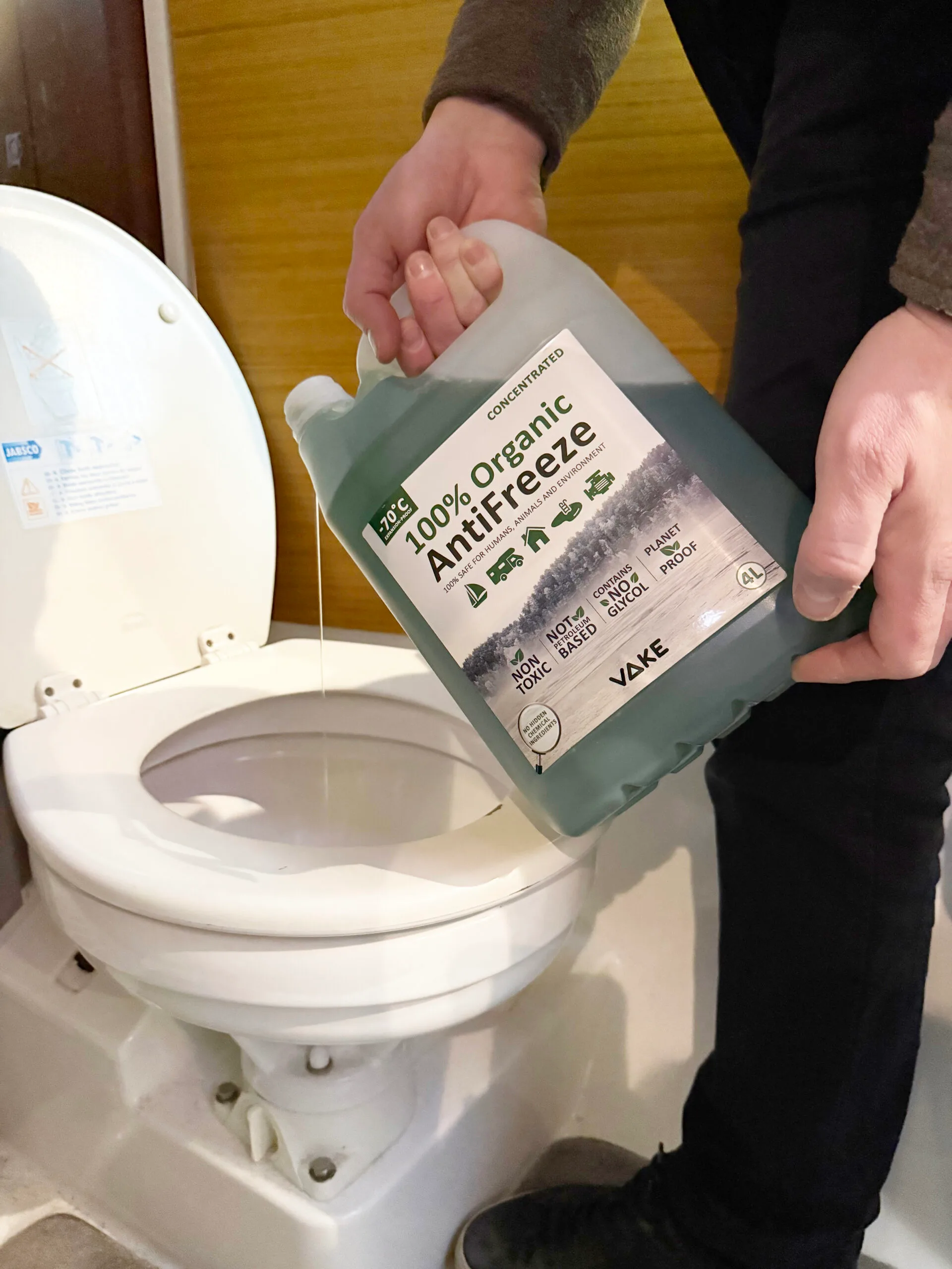 A person pouring VAKE 100% Organic AntiFreeze from a 4-liter container into a toilet in a boat bathroom, demonstrating how to protect the plumbing system from freezing.